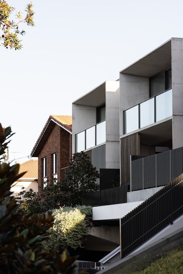 Exterior view of a contemporary multi-level residential building on a sloped site. The facade combines exposed grey concrete, dark vertical metal screening, glass balcony balustrades, and recessed upper-level terraces. Adjacent brick and terracotta-tiled homes are visible, with landscaped planting in the foreground and a prominent external stairway descending along the frontage.