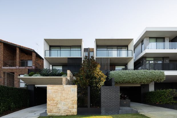 Street-facing view of a contemporary multi-unit residential development with three adjoining modern dwellings. The buildings feature flat rooflines, projecting upper-level volumes, large glazed openings, dark vertical screening, balconies, landscaped hedges, and contrasting pale stone and dark brick boundary walls. A traditional brick dwelling is visible at the left edge.