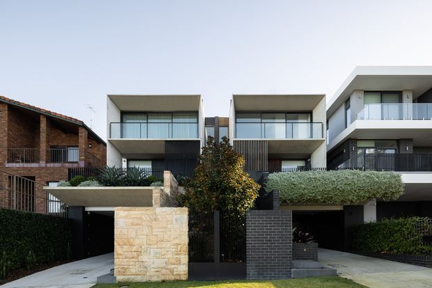 Street-facing view of a row of contemporary multi-storey residences with strong geometric forms, projecting box-like upper levels, extensive glazing, balconies, dark vertical screening, and integrated garages. The homes combine pale rendered surfaces, dark brickwork, stone garden walls, clipped hedges, and narrow landscaped entries. A contrasting older brick house is visible at the left edge.