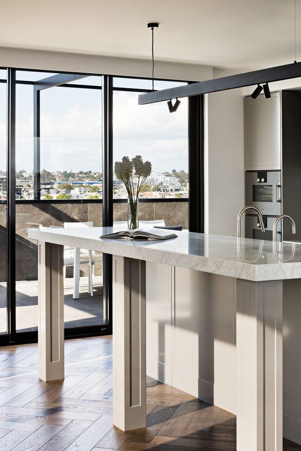 Bright contemporary kitchen with a large white stone island featuring paneled cabinetry, integrated sink and curved brushed-metal tapware. Full-height black-framed glazing opens to a balcony or terrace with urban views. Warm timber parquet flooring, dark window frames, integrated appliances and a linear black pendant/track light create a refined, modern interior.