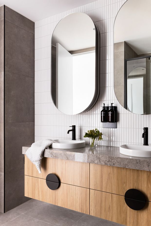 A contemporary bathroom vanity with a double-basin arrangement, light timber floating cabinetry, a grey stone benchtop, black tapware and hardware, and rounded vertical mirrors. The walls combine slim white vertical tiles with large dark grey tiles, while the floor is finished in matching grey tile. The space has a clean, refined and warm modern appearance.