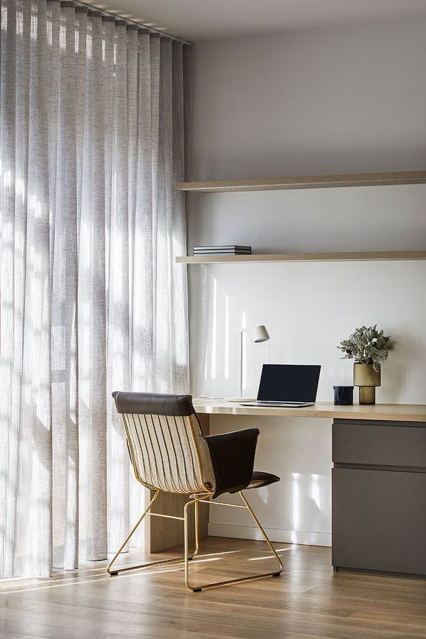 A bright, minimalist home office with a built-in timber desk and floating timber shelves against a white wall. Sheer floor-to-ceiling curtains filter sunlight at the window, while a dark upholstered and brass-framed chair, laptop, desk lamp and small plant complete the workspace. The room has warm timber flooring and dark grey built-in cabinetry.
