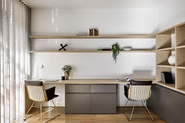 A bright, carefully designed home office with a long built-in timber desk, central dark cabinetry, open display shelving, and two upholstered lounge-style chairs. Full-height sheer curtains provide soft natural light, while potted plants and minimal decor add warmth to the clean, contemporary space.