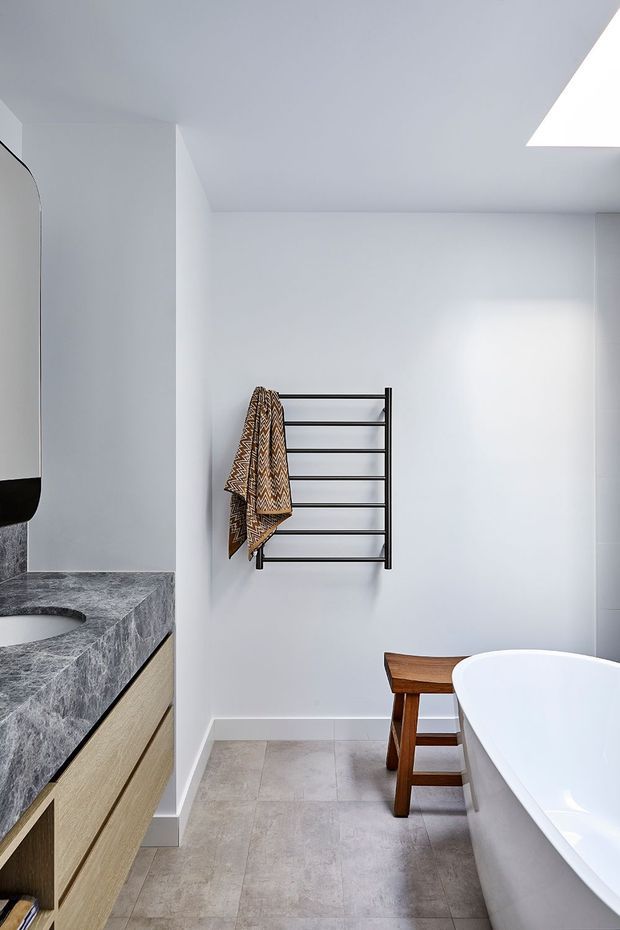 Bright, minimalist bathroom with white walls and ceiling, a skylight, pale stone-look floor tiles, a freestanding white bathtub, timber stool, wall-mounted black heated towel rail, and a timber vanity with a dark grey stone benchtop and integrated basin. A partial mirror is visible at left.