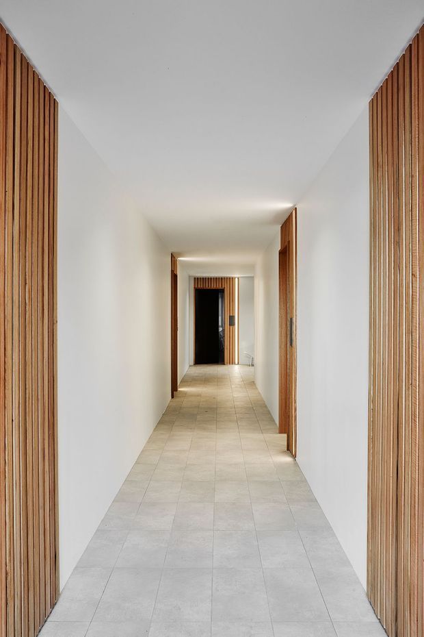 A long, narrow minimalist hallway with smooth white walls and ceiling, pale square tiled flooring, and warm timber vertical slat screens framing both sides. Timber door frames and doors line the corridor, with recessed ceiling lighting creating a clean, calm, symmetrical composition.