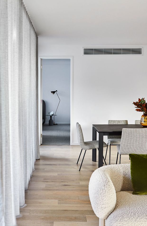 A bright, refined open-plan interior featuring a dining area and partial living room. Full-height sheer curtains line the left side, while pale timber flooring, white walls, a dark rectangular dining table, textured upholstered dining chairs, and a curved cream boucle armchair create a calm contemporary setting. A doorway leads to a carpeted room with a floor lamp, and a linear air-conditioning vent is integrated into the wall above the dining area.