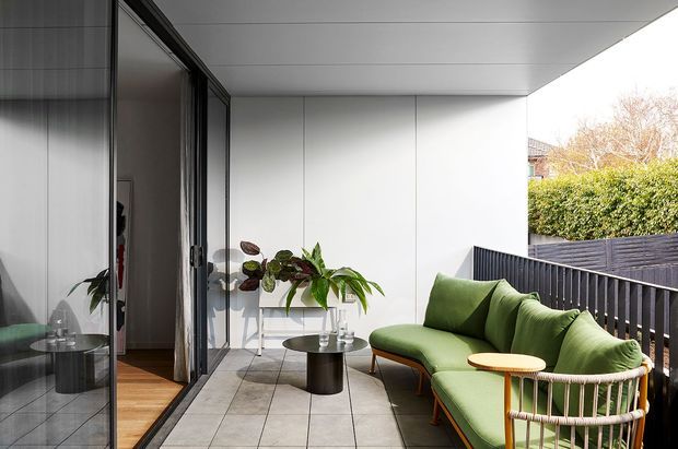 A contemporary covered outdoor terrace or balcony with a green upholstered sofa, round timber side table, dark cylindrical coffee table, potted plants, and a black vertical-slat balustrade. Large sliding glass doors connect the terrace to the interior, where timber flooring and reflected furnishings are visible. The space has a clean, minimalist architectural shell with pale wall and ceiling panels and grey tiled flooring.