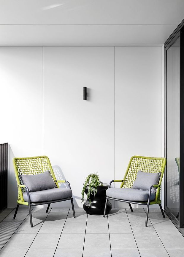 A minimalist seating area with two lime-green woven lounge chairs, grey cushions, and black metal frames arranged around a black planter. The space has large light-grey tiled flooring, white paneled walls, a simple wall light, and dark-framed glazing on the right. The composition is bright, clean, and contemporary.
