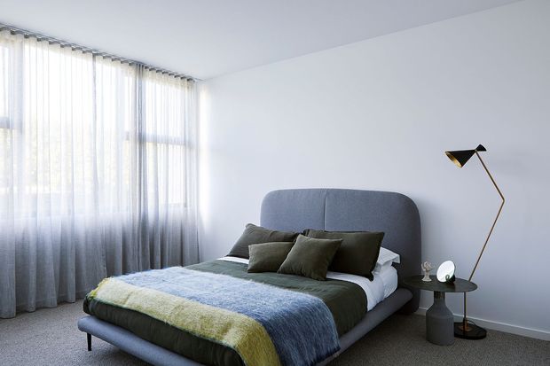 A calm, contemporary bedroom with a low upholstered bed, rounded grey headboard, layered green and blue bedding, and neutral cushions. Full-height sheer curtains filter soft daylight from a large window, while a slim black-and-brass floor lamp and compact side table provide refined accents against pale walls and carpeted flooring.