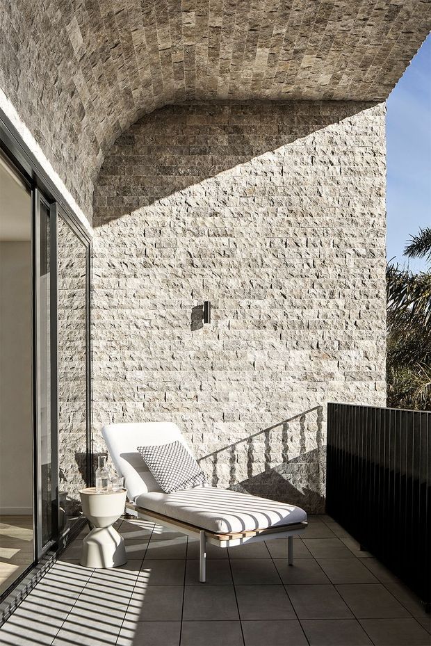 A sunlit contemporary outdoor terrace or balcony with a white stone masonry wall and vaulted stone soffit. Large dark-framed sliding glass doors open to the interior, while a white upholstered chaise lounge and small sculptural side table sit on large grey tiled flooring. A dark vertical-slat privacy screen and strong architectural shadows add contrast.