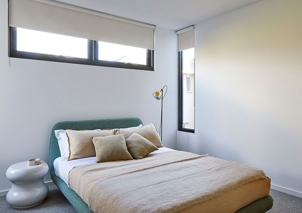 A bright, minimalist bedroom with a teal upholstered bed, layered neutral linen bedding and cushions, a sculptural white side table, and a slim brass-toned floor lamp. White walls, grey carpet, and high horizontal windows with roller blinds create a clean contemporary interior.