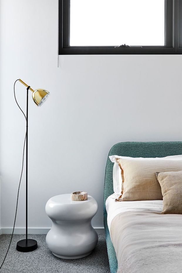 A softly styled bedroom corner with a partially visible bed, textured teal upholstered headboard, layered cream and beige bedding, a sculptural white bedside table, and a slim black-and-brass floor lamp. The room has pale grey walls, grey carpet, and a black-framed upper window, creating a clean, refined contemporary look.