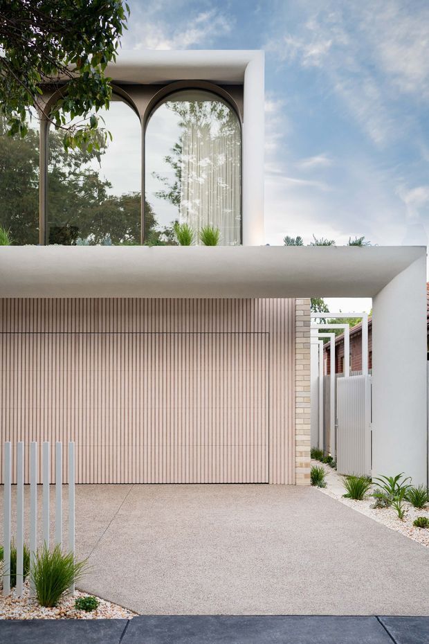 A contemporary residential front facade viewed from the street, featuring a white rendered upper volume with large arched glazed windows, a broad projecting canopy, and a pale vertically ribbed garage door. A textured concrete driveway leads to the garage, with white gravel and sculptural grasses along the narrow side garden. The composition uses strong geometric lines, arches, and a restrained neutral palette.