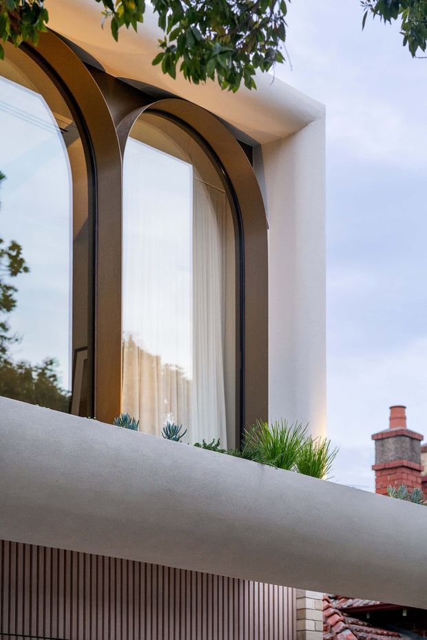 Close-up exterior view of a refined contemporary residential facade, featuring tall arched bronze-toned framed windows set into smooth white stucco, a projecting curved balcony edge, and vertical timber-toned screening below. Small planted greenery sits on the balcony, with a neighboring brick chimney and tiled roof visible in the background.