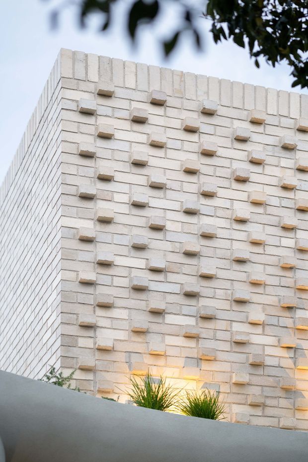 Close-up architectural exterior view of a pale cream-white brick wall with projecting bricks arranged in a regular decorative pattern. The wall forms a clean modern corner, with warm lighting and ornamental grasses visible at the base, partially framed by blurred foliage.