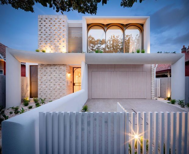 A contemporary two-storey residential facade viewed from the street at dusk. The home features smooth white rendered walls, pale textured brickwork, a large upper-level window with repeated arched divisions, horizontal screening, a deeply recessed double garage with vertical slatted detailing, and a white vertical-slat front fence. Low-maintenance planting lines the entry and driveway.