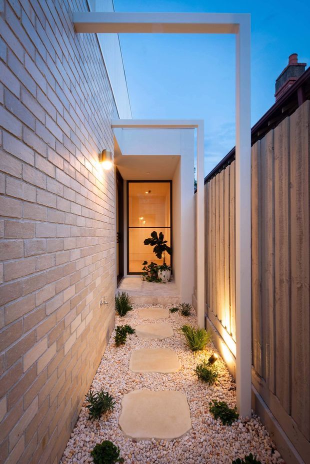A narrow landscaped side passage leading to a glazed entry door, bordered by a pale exposed-brick wall on one side and a tall vertical timber fence on the other. White rectangular overhead frames create a covered architectural walkway, while warm wall and ground lighting illuminates pale gravel, stepping stones, and small drought-tolerant plantings. The scene is photographed at dusk with a contemporary minimalist feel.