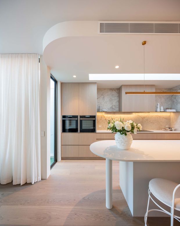 Bright, refined contemporary kitchen with pale timber cabinetry, integrated black appliances, a marble-look stone backsplash and benchtop, and a rounded white island with curved supports. The space features pale timber flooring, a large skylight, concealed warm under-cabinet lighting, full-height glazing with sheer curtains, and softly curved ceiling and wall details.