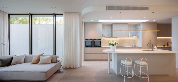 Bright open-plan contemporary kitchen and living area. A pale grey sectional sofa sits beside a light timber kitchen with integrated appliances, a large pale stone island, slim open shelving, warm under-cabinet lighting, and white bar stools. Full-height black-framed glazing and sheer curtains bring in natural light, while a curved ceiling transition adds architectural softness.