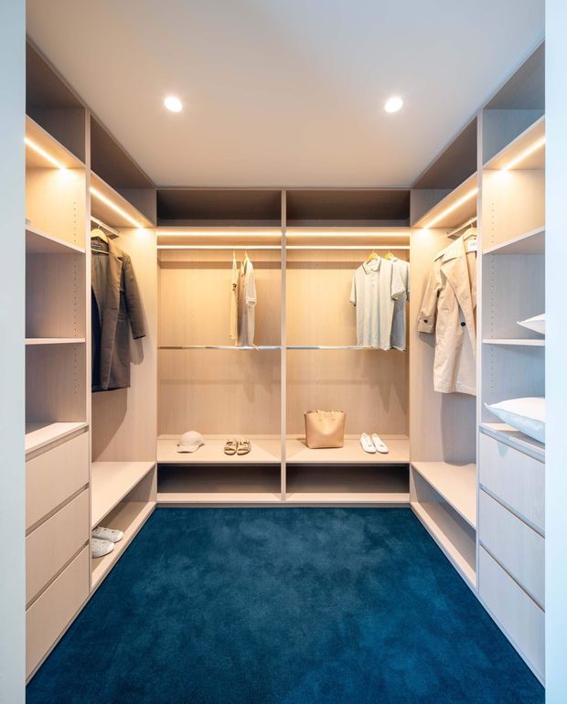 A compact, well-organised walk-in wardrobe with pale timber-look built-in cabinetry, open hanging rails, drawers, shelving, integrated warm LED strip lighting, and a deep blue carpeted floor. Clothing, shoes, folded linens, and accessories are displayed within the joinery. The space has a clean contemporary fitted-out appearance.