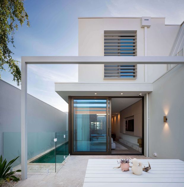 A modern white two-storey residence viewed from an outdoor terrace, with a projecting flat-roofed upper level, horizontal louvred windows, frameless glass pool fencing, and a glazed sliding door opening into a contemporary living area. The terrace includes a white dining table, while a narrow plunge pool runs alongside the house.