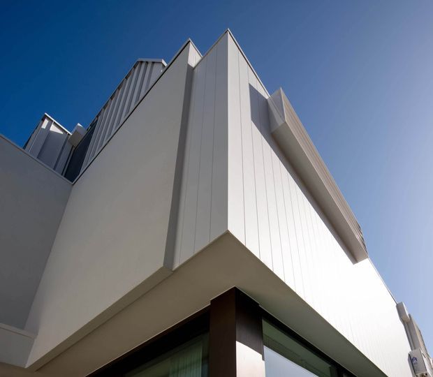 Low-angle architectural photograph of a contemporary multi-storey building facade against a clear blue sky. The composition emphasizes sharp angular projections, folded roofline forms, vertical ribbed light-coloured cladding, dark structural accents, and large glazed openings at the lower level.