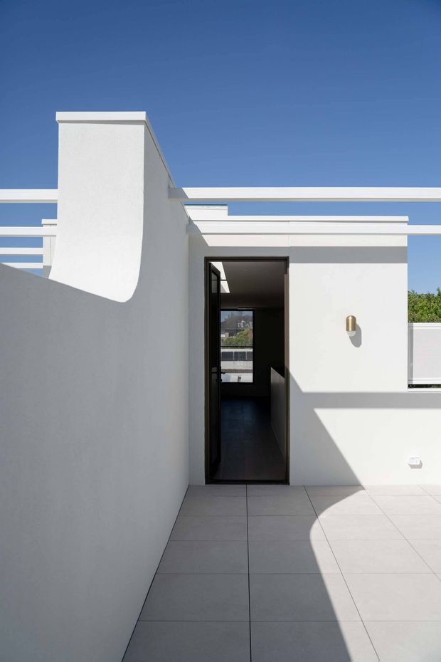A bright, minimalist rooftop terrace or courtyard with white rendered walls, a flat roofline, geometric parapets and large pale square floor tiles. A dark-framed doorway opens into the building, while strong sunlight creates angular shadows across the terrace. The architecture has clean modern lines against a clear blue sky.