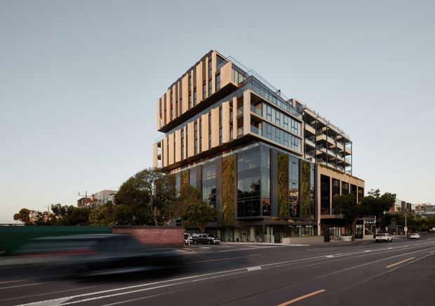 Street-level exterior view of a contemporary multi-storey mixed-use or commercial building with a stepped, cantilevered massing. The facade combines extensive glazing, warm vertical timber-toned fins, dark structural framing, balconies, and landscaped vertical greenery. The building fronts a broad urban roadway with surrounding trees and low-rise context.