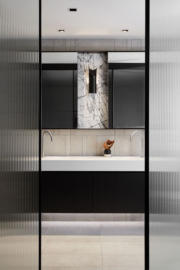 A highly designed contemporary bathroom vanity viewed through fluted translucent glass panels. The room features a long white benchtop with two curved chrome-silver wall-mounted faucets, pale large-format tiled walls and floor, dark vertically fluted cabinetry, and a central dramatic marble feature panel with an integrated dark cylindrical wall light. Recessed ceiling lights and strong black framing create a refined, high-contrast composition.