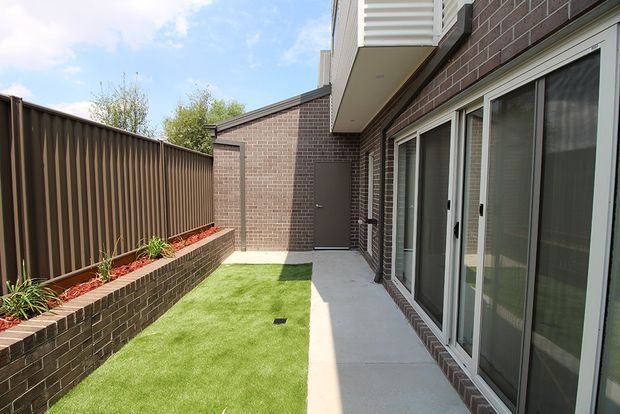 A narrow landscaped courtyard or side passage beside a modern brick building, featuring a small artificial grass lawn, raised brick planter with red mulch and plants, tall brown privacy fencing, and a concrete walkway along full-height glazed doors and windows. The scene is bright and contemporary, with an upper-level projecting volume visible above.