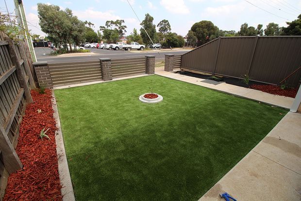 A compact, neatly landscaped residential backyard with a rectangular artificial grass lawn, a small circular fire pit or garden feature at its centre, red mulch garden beds, and dark horizontal privacy fencing. Concrete paving borders the lawn, with a gate opening toward a suburban street and parked cars beyond.