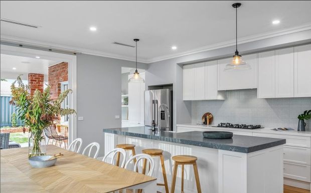 Bright contemporary kitchen and dining area with white shaker-style cabinetry, a large grey stone island with seating, stainless-steel appliances, pale tiled splashback, pendant lighting, and a timber dining table. Sliding glazed doors open toward an outdoor area, with exposed brickwork visible near the opening.