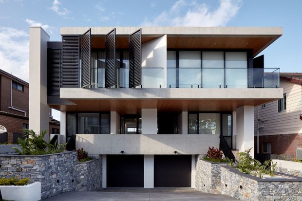 Front exterior view of a contemporary multi-storey residence with a strongly horizontal composition, projecting flat roof and balconies, white rendered walls, exposed concrete elements, dark aluminium-framed glazing, louvred screens, and warm timber soffits. The house includes a lower-level double garage, stone retaining walls, landscaped planting, and a driveway.