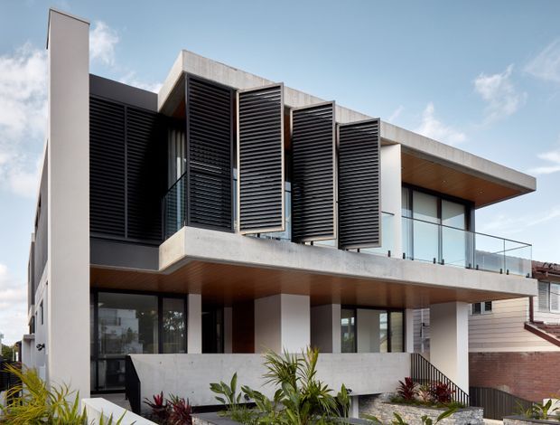 Exterior architectural photograph of a contemporary multi-level residence. The facade combines exposed concrete slabs and walls, dark vertical louvred screens, extensive glazed openings with clear glass balcony balustrades, and warm timber soffits. Tropical landscaping and neighbouring residential buildings are visible around the property under a bright sky.