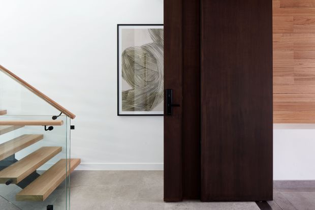 A refined interior view of a stair landing or hallway, featuring a floating timber staircase with clear glass balustrades and a timber handrail on the left. A large dark timber pivot door or wall panel dominates the centre, with a black lever handle. The background has white walls, a framed abstract artwork, light neutral flooring, and warm timber wall cladding visible at upper right.
