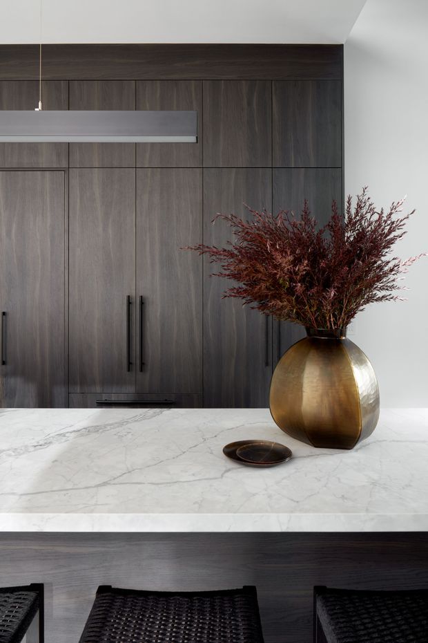 A refined contemporary kitchen focused on a dark timber island and full-height dark wood cabinetry. The island has a bright white marble-look stone benchtop, with black woven bar stools beneath a slim linear pendant light. A large bronze-toned vase with dried branches provides a warm decorative accent against the otherwise monochromatic palette.