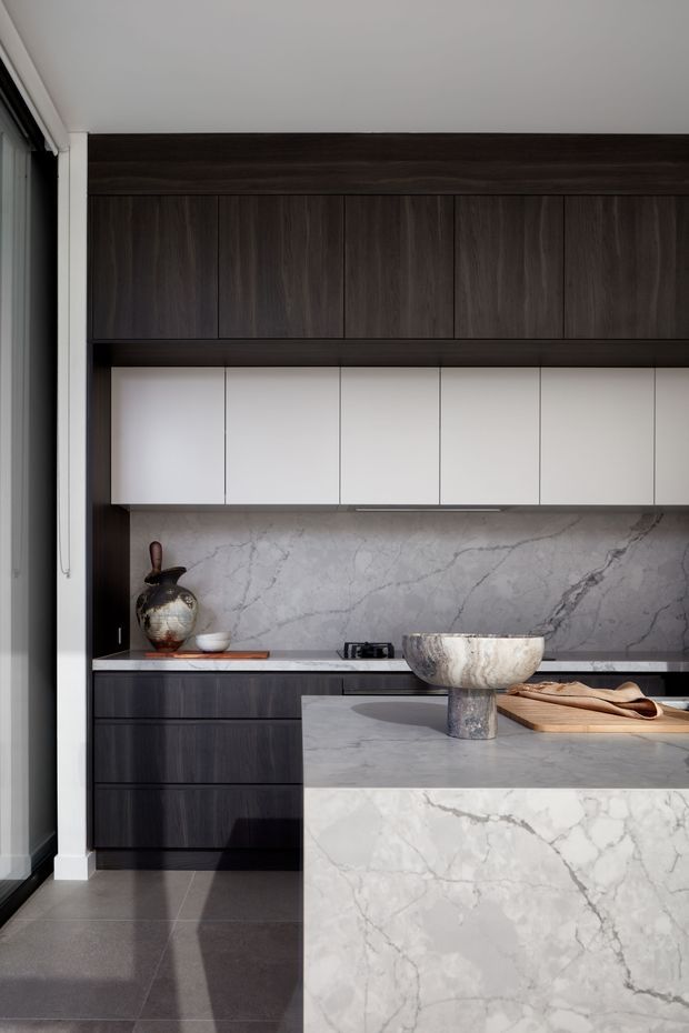 A refined modern kitchen with a dark timber-grain cabinetry wall, crisp white overhead cupboards, and dramatic white veined stone used for the splashback, benchtops, and waterfall island. The space has a restrained monochromatic palette with warm timber accessories and soft natural light.
