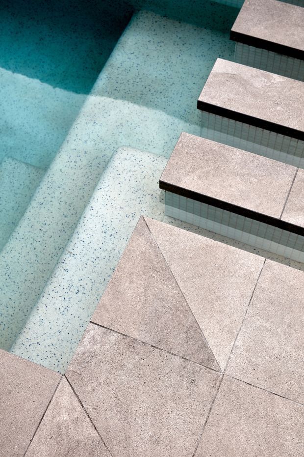 Close-up architectural detail of a swimming pool with pale blue speckled interior surfaces and a series of light grey-beige tiled steps or platforms. The pool edging and platforms use large-format textured stone-look tiles with dark grout lines, while the vertical risers are clad in small pale blue mosaic tiles. Strong sunlight creates geometric shadows across the water and surfaces.