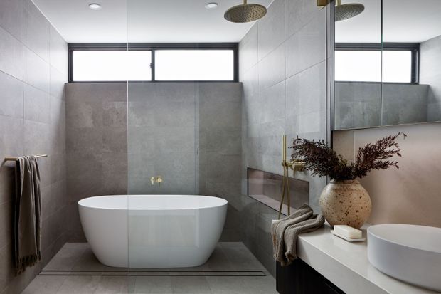 A contemporary bathroom featuring a freestanding oval bathtub behind a frameless glass shower screen, large grey concrete-look wall and floor tiles, a wall-mounted rainfall shower, clerestory windows with black frames, and a white vanity with an integrated basin. A large mirror reflects the shower area, while neutral textiles and a ceramic vase add warmth.