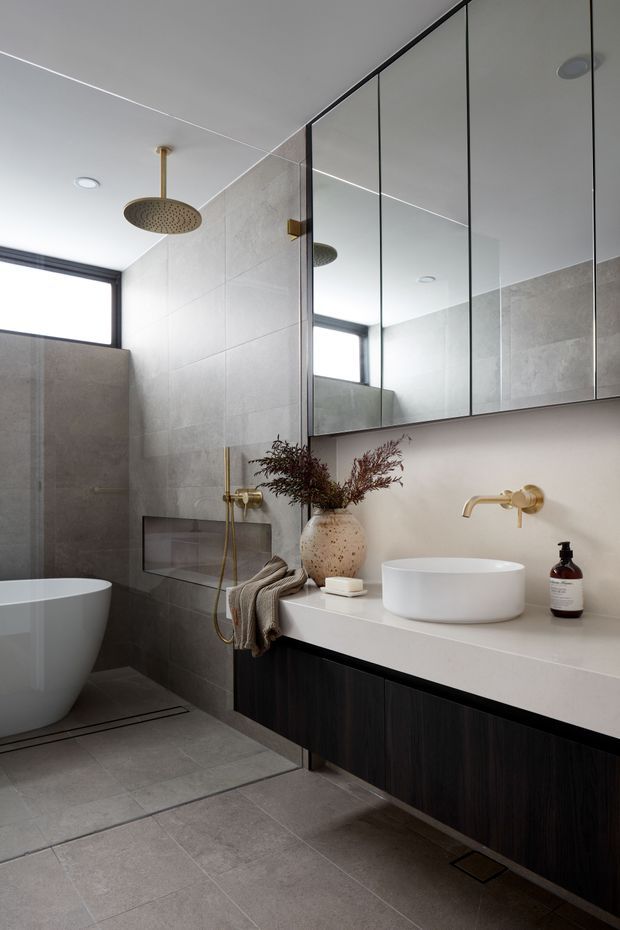 Contemporary bathroom with large-format grey wall and floor tiles, a freestanding white bathtub, walk-in shower with rainfall showerhead, floating dark timber vanity, white stone benchtop, vessel basin, wall-mounted brass tapware and mirrored cabinet. A high horizontal window provides natural light, while warm brass fittings and muted styling add refinement.