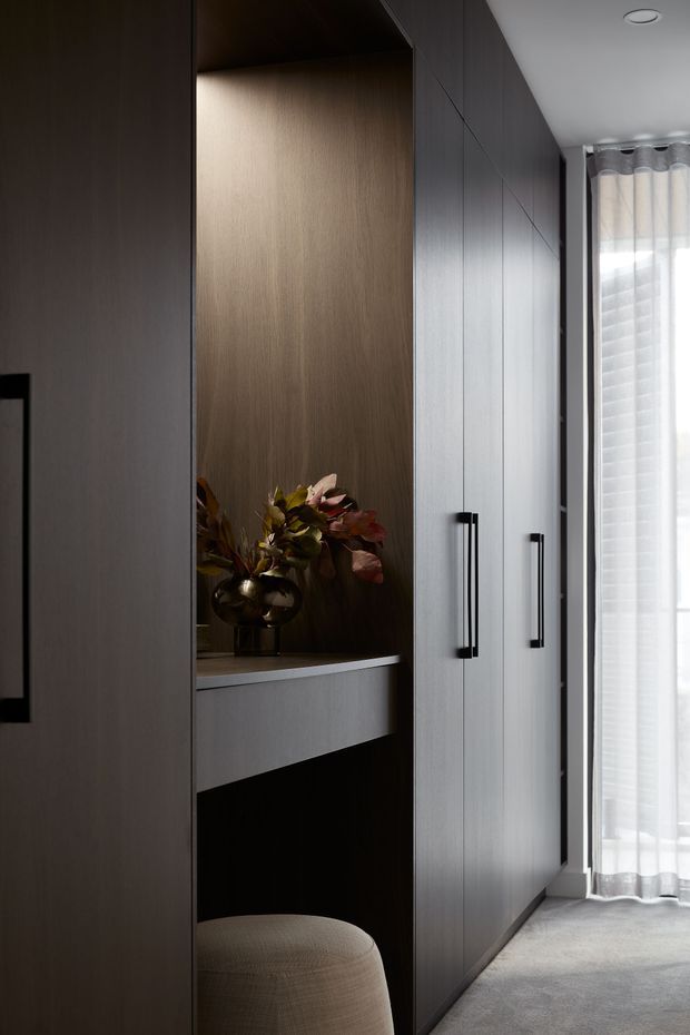 A moody, refined interior view of a built-in wall of dark timber-toned cabinetry with long black handles. An illuminated recessed display niche contains a decorative vase and foliage above a floating or built-in desk surface, with a rounded upholstered stool beneath. Sheer curtains and a bright window are visible at the end of the space, while carpet flooring and a smooth white ceiling complete the setting.