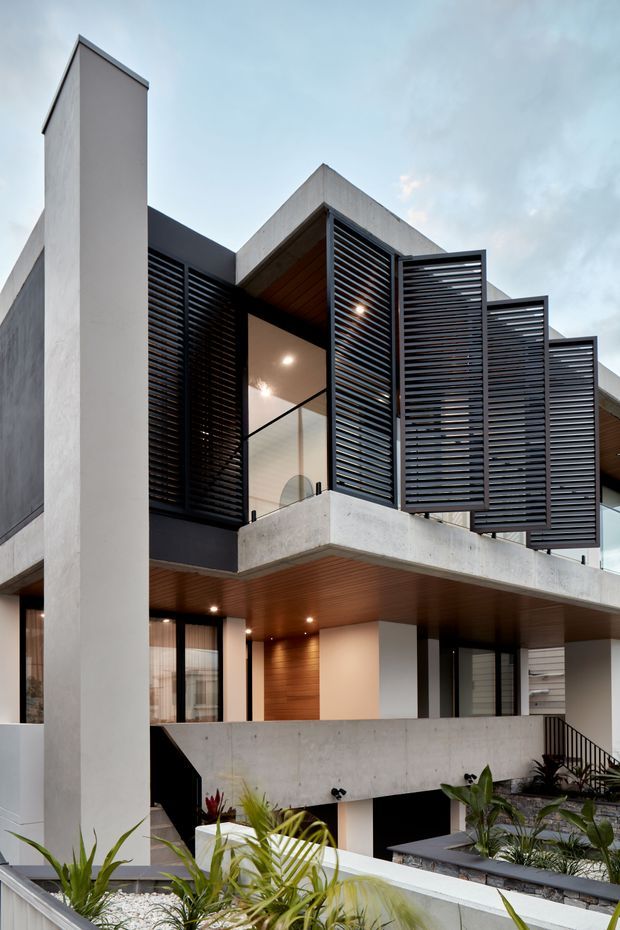 A contemporary multi-level residential facade viewed from a landscaped courtyard. The building features raw concrete volumes and balconies, dark horizontal louvred screens, extensive glazing, black metal framing, and warm timber-lined soffits. A tall vertical concrete wall anchors the composition, while planters and a reflecting-water feature occupy the foreground.