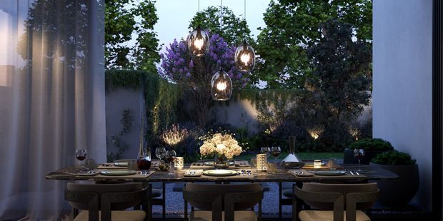 An elegantly styled dining area opens onto a landscaped courtyard or garden through large glazed openings. A long dark dining table is set for dinner with tableware and wine glasses, surrounded by upholstered dining chairs. Three sculptural pendant lights hang above the table, while warm integrated lighting highlights the planting and textured garden wall beyond. The scene has a refined, intimate evening atmosphere.