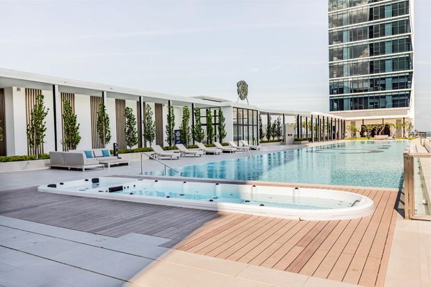 A bright rooftop or elevated pool area with a long rectangular swimming pool and an attached spa set into a broad timber-look deck. White poolside loungers, vertical planted screens, glass balustrades, and a sleek low-rise pavilion frame the space, with a modern high-rise building visible in the background.