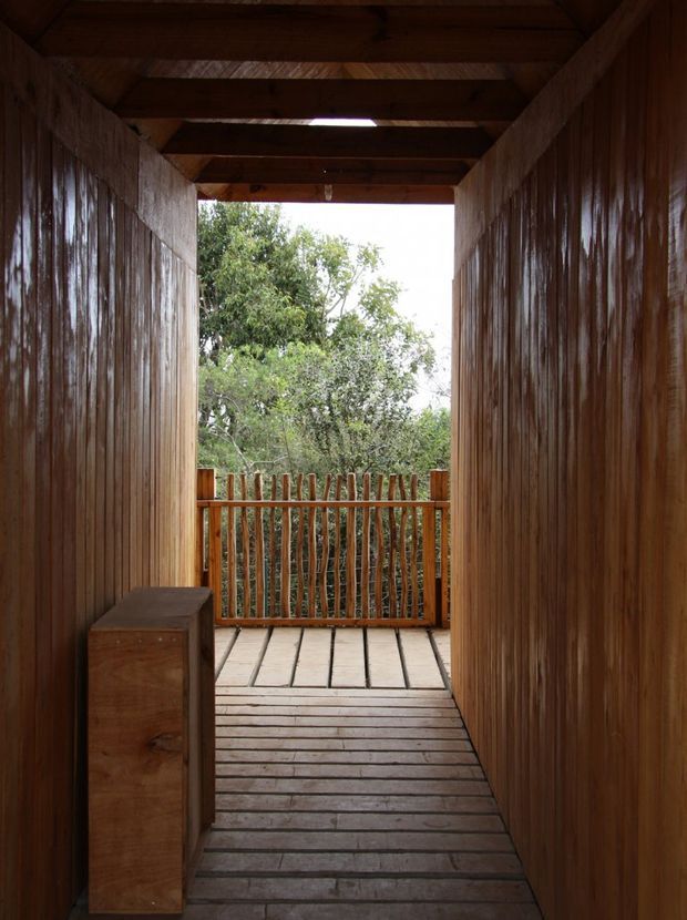 A narrow, enclosed timber passage leads out to a small elevated wooden deck or balcony surrounded by trees. The passage has richly stained vertical timber wall cladding, exposed horizontal timber roof beams, timber plank flooring, and a rustic branch-style timber balustrade. A simple built-in timber cabinet or bench sits along the left wall. The scene is warm, natural, and heavily wood-lined.