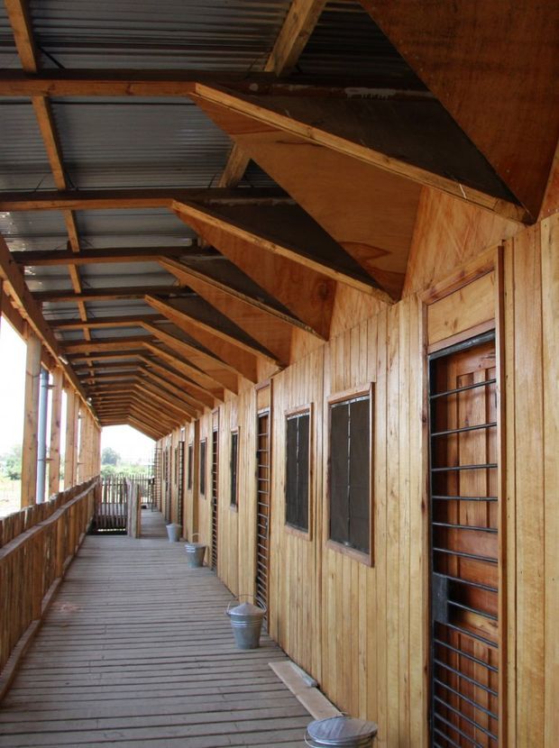 A long, covered exterior timber walkway or veranda running alongside a timber-clad building. The corridor has a timber plank floor, exposed timber roof trusses, corrugated metal roofing, repeated timber-framed doors and windows with dark security grilles, and several metal buckets placed along the wall. Bright rural landscape is visible beyond the open side.