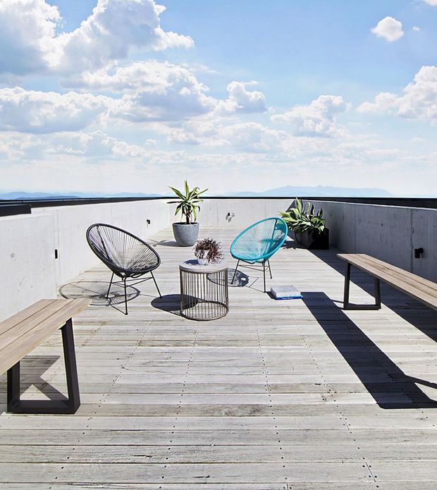 A spacious rooftop terrace or deck with pale weathered timber decking, low smooth concrete parapet walls, and minimalist outdoor furniture. Two wire-frame lounge chairs, a small round wire table, and long simple timber benches are arranged against a broad sky and distant mountain horizon. Potted plants add greenery, while strong sunlight creates crisp shadows across the deck.