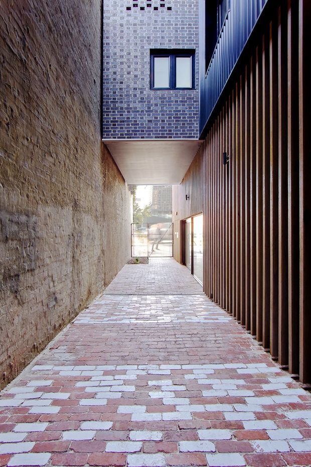 A narrow urban passageway between buildings, with a reclaimed brick-paved ground plane, a rough exposed masonry wall on the left, and a tall dark timber or metal vertical-screen facade on the right. A cantilevered upper level with pale grey brickwork and a small window spans overhead, creating a covered section. The passage opens toward a gated street-facing area and distant urban buildings.