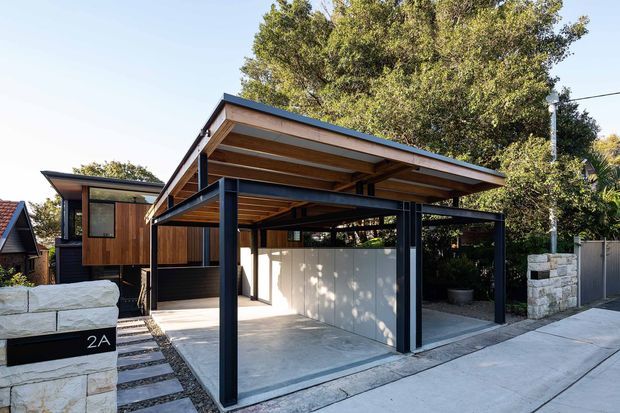 Street-facing exterior view of a contemporary house with a prominent open-sided carport. The carport has a broad sloping roof with exposed timber soffit and dark metal posts and beams, alongside a timber-clad upper volume and dark-framed windows. A concrete driveway, stepping-stone path, low pale masonry wall, and established trees complete the frontage.