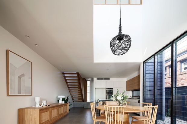 Bright open-plan dining and kitchen area with a timber dining table and spindle-back chairs, a timber sideboard, light neutral walls, dark flooring, and extensive black-framed glazing. A sculptural woven pendant hangs beneath a large skylight, while an open timber staircase leads toward the rear of the home. The interior has a clean contemporary feel with warm natural timber accents.